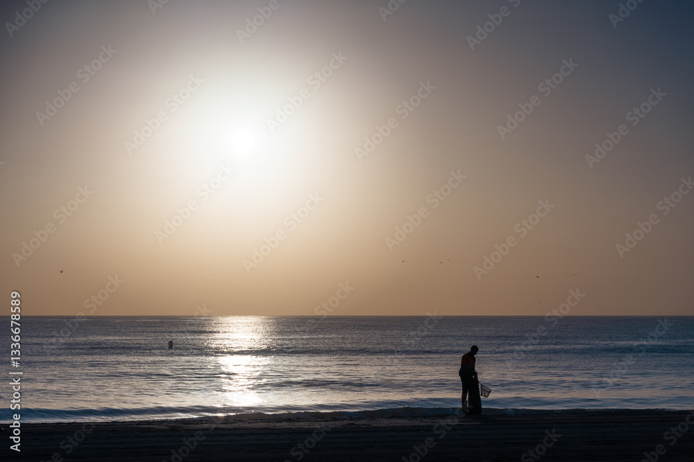 silhouette of a man with a net cleaning the sand on the beach near the seashore at dawn silhouette of a man with a net cleaning the sand on the beach near the seashore at dawn