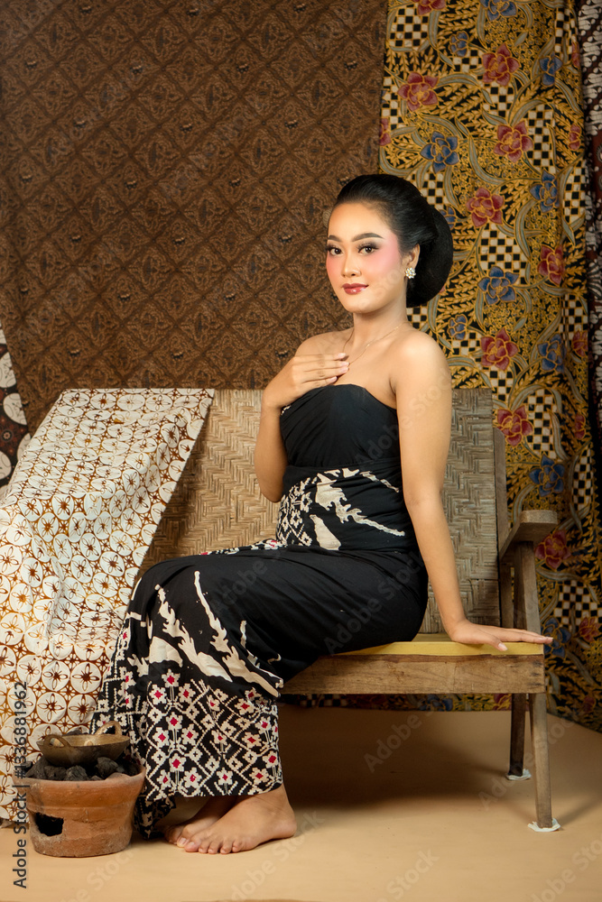 Fototapeta premium Elegant Indonesian woman wearing traditional batik fabric, sitting on a wooden bench. Vintage ethnic backdrop with rich patterns, cultural heritage, and graceful beauty in classic style.