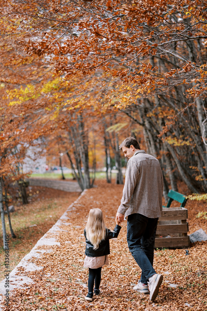 Fototapeta premium Dad looks at the little girl as he walks with her holding hands along a path in the autumn forest. Back view