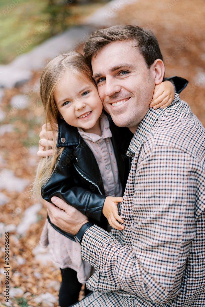 Obraz premium Smiling little girl hugs her dad neck as he squats in an autumn park