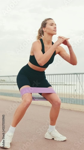 Vertical video, Young woman doing squats with resistance band on a promenade. She is wearing black top and shorts. Ocean view and palm trees. Cloudy sky. photo