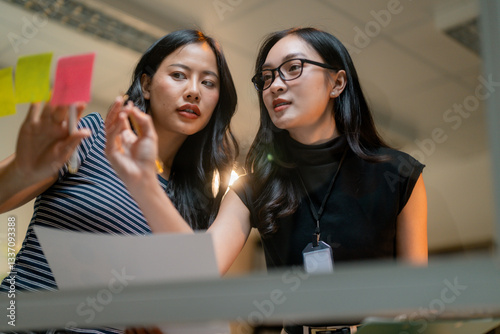 Businesswomen analyzing project planning using sticky notes on glass wall in the office