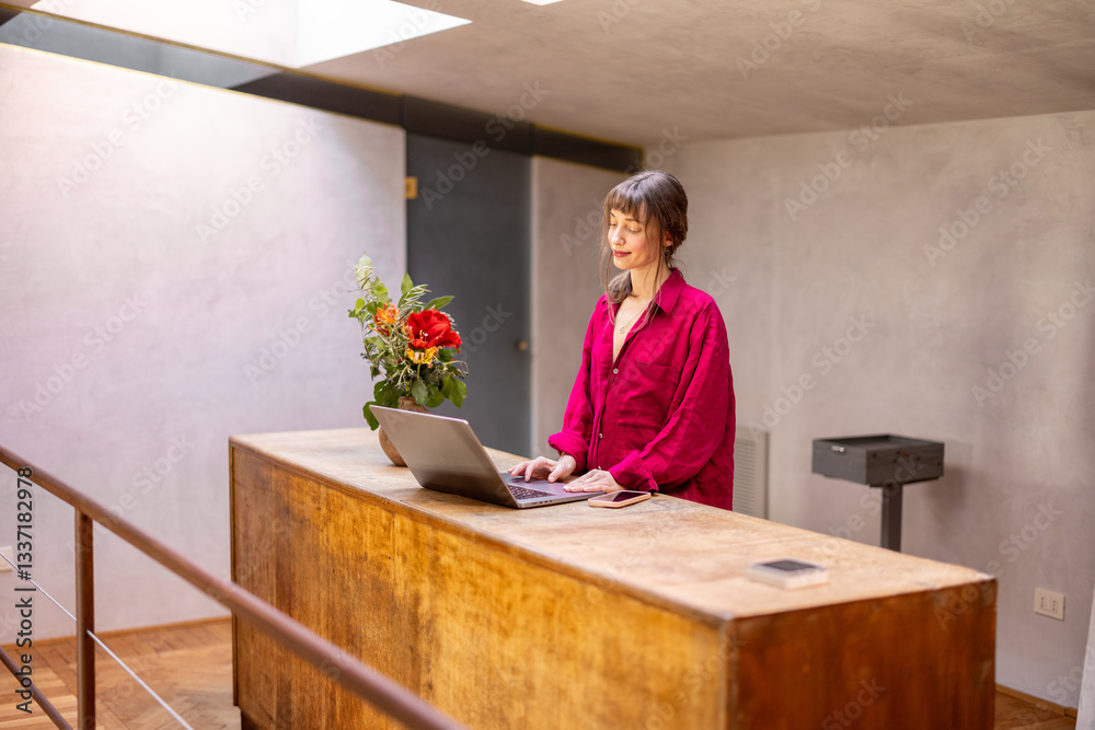 A stylish woman in red leans over a wooden desk, engaged in work on her laptop. The setting embodies focus, creativity, and modern remote work culture in a cozy, minimalist home environment