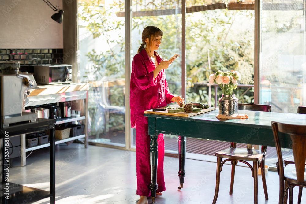 A woman in soft pajamas enjoys a peaceful morning in her sunlit kitchen, checking a recipe on her phone while preparing fresh vegetables. The scene embodies slow living and mindful routines