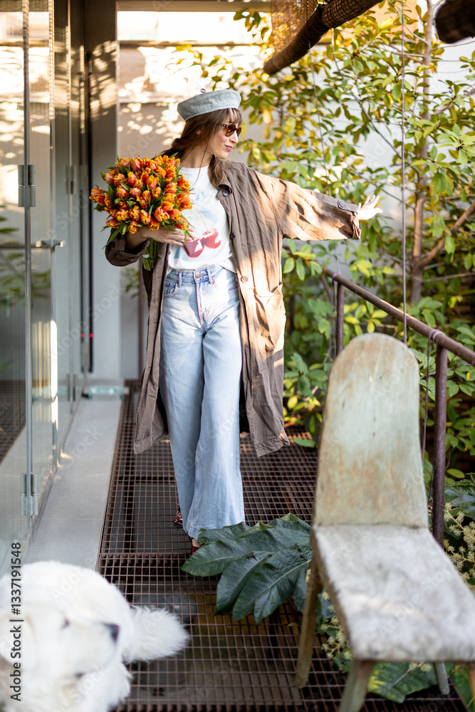 Fashionable woman in sunglasses and a beret sits on a patio with a bouquet of vibrant tulips and a large white dog. The modern setting, warm sunlight, and cozy atmosphere create elegant moment