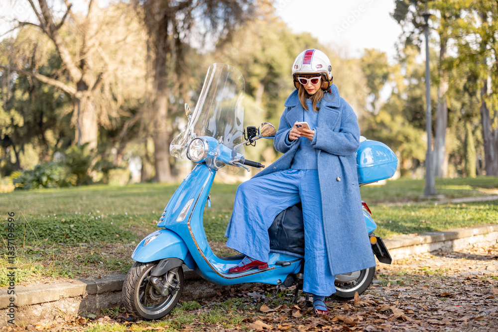 A stylish woman in a blue coat and helmet leans on her scooter while checking her phone in a peaceful park. Fashion, independence, and leisure in an urban nature setting