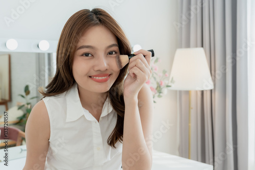 Asian woman holding makeup brush gently against cheek while smiling wearing white sleeveless top. Radiant complexion with warm friendly expression. Beauty concept focusing on skincare and cosmetics.