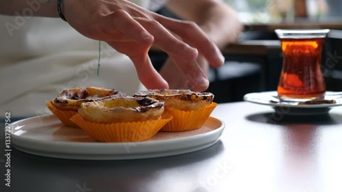 Close-Up of a Hand Picking a Dessert from a Plate Next to Tea on a Café Table with Blurred Crowd in Background