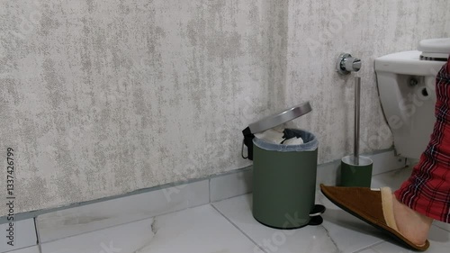 Person Throws Tissue into Pedal Trash Bin Next to Toilet in Marble Bathroom While Wearing Pajamas and Slippers