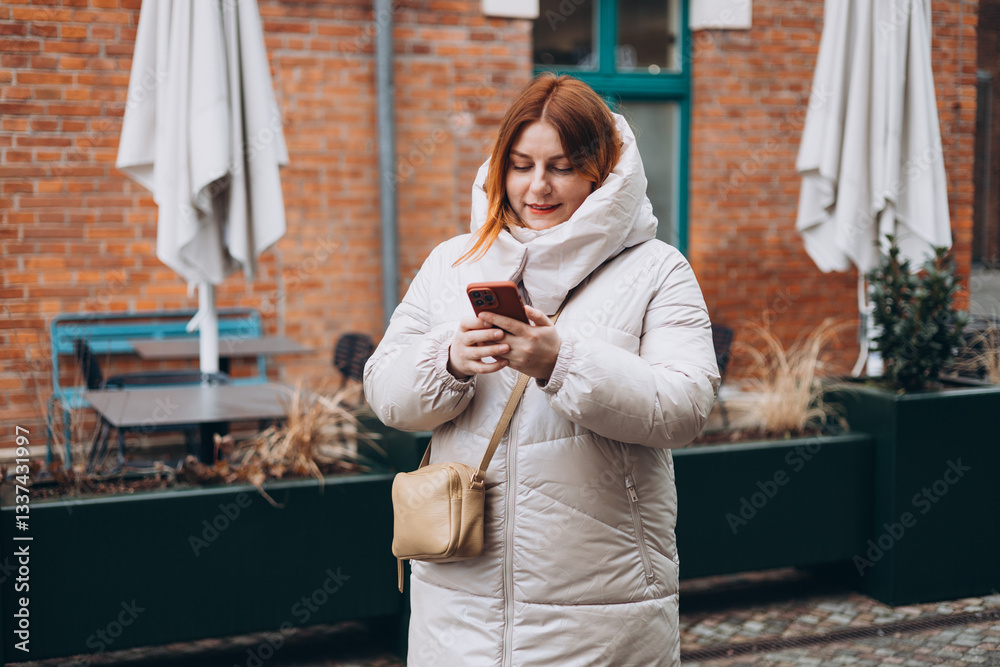 Young stylish 30s woman writes message on smartphone walking on city street. Urban lifestyle concept. Autumn time. Charming female wearing stylish outfit chatting online. High quality photo.