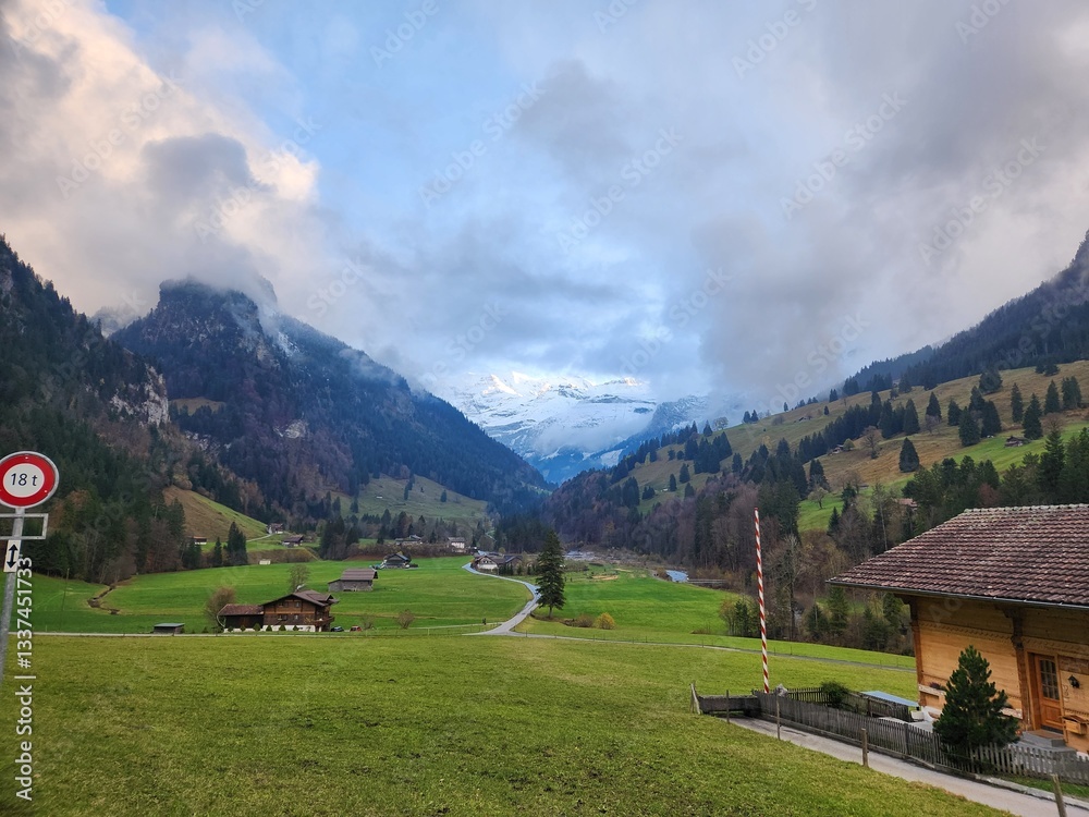 Obraz premium Idyllic Swiss Mountain Village: A Serene Landscape of Rolling Green Hills, Charming Wooden Chalets, and Snow-Capped Peaks Under a Dramatic Cloudy Sky
