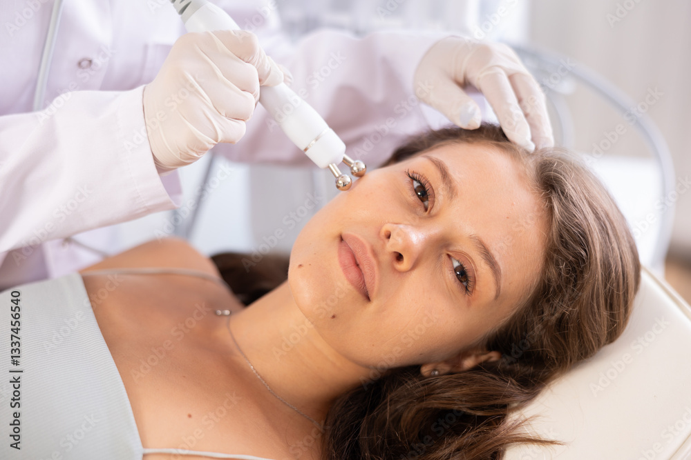 Naklejka premium Positive young girl undergoing radiofrequency skin tightening procedure enhancing facial skin tone and preventing wrinkles. Modern technologies in cosmetology and aesthetic medicine