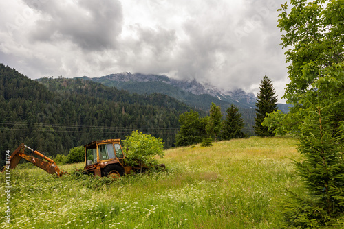 Obraz na płótnie vista panoramica su un ambiente naturale di montagna nell'Italia settentrionale, di giorno, in estate, con cielo nuvoloso e coperto e con una vecchia ruspa abbandonata in un campo con erba alta w salonie