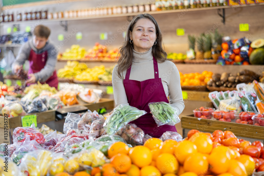 Young woman seller in apron lays out green peas on counter in vegetable shop Young woman seller in apron lays out green peas on counter in vegetable shop