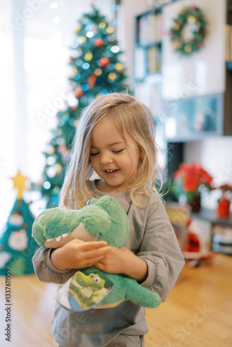 Little smiling girl hugging soft toy crocodile while standing near Christmas tree with gifts photo