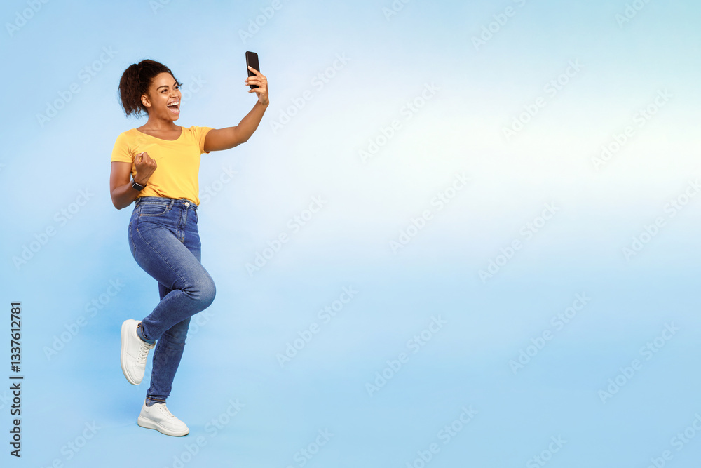 A cheerful African American woman stands against a light blue background, smiling as she captures a selfie with her smartphone while wearing casual attire and stylish sneakers, copy space
