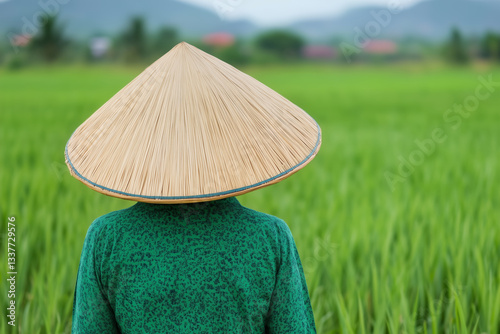Asian farmer wearing a traditional conical straw hat standing in a lush green rice field, rural agricultural scene with blurred mountains and village in the background