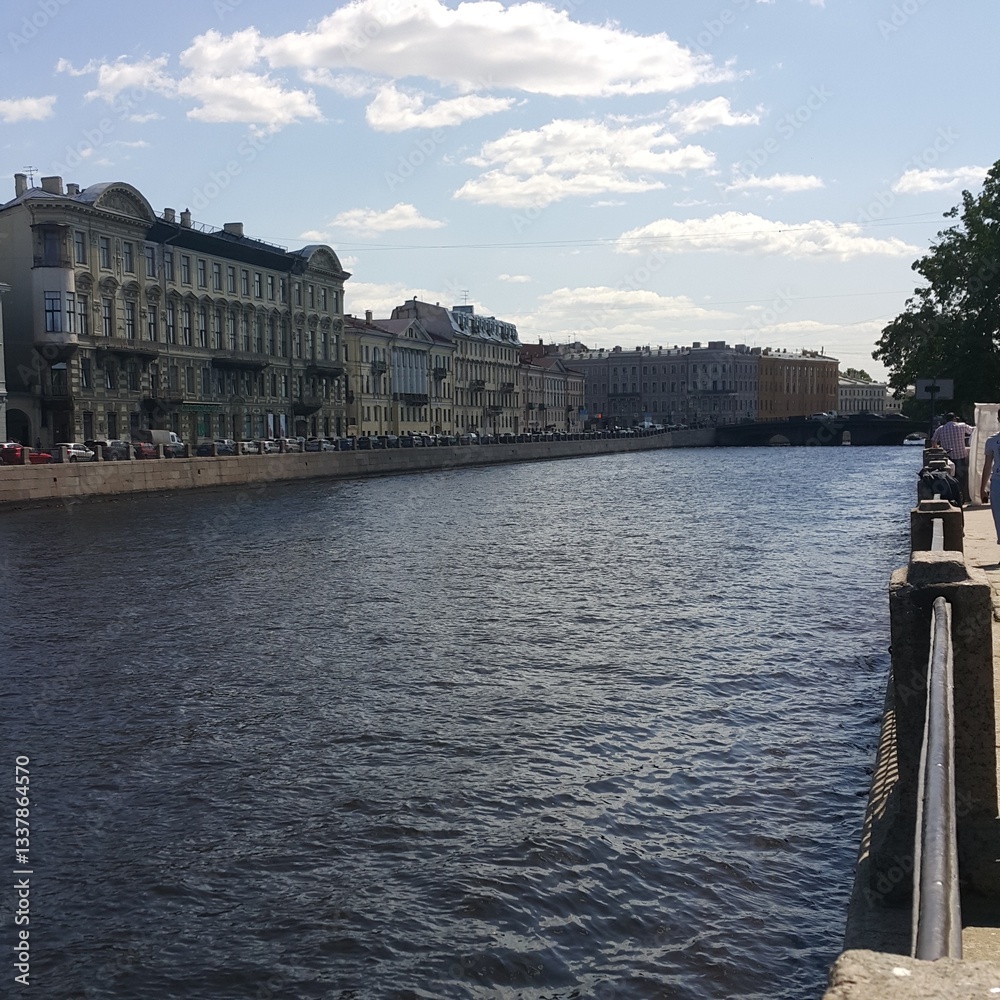 Fototapeta premium View of Neva River embankment in Saint Petersburg with historic buildings under a partly cloudy sky. Captured in daylight displaying reflections in the water and architecture along the canal. 