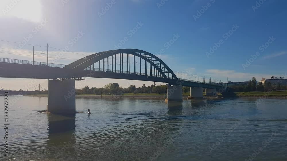 View of the tranquil Sava River in Belgrade, with cars crossing the historic Old Sava Bridge beneath a clear blue sky View of the tranquil Sava River in Belgrade, with cars crossing the historic Old Sava Bridge beneath a clear blue sky
