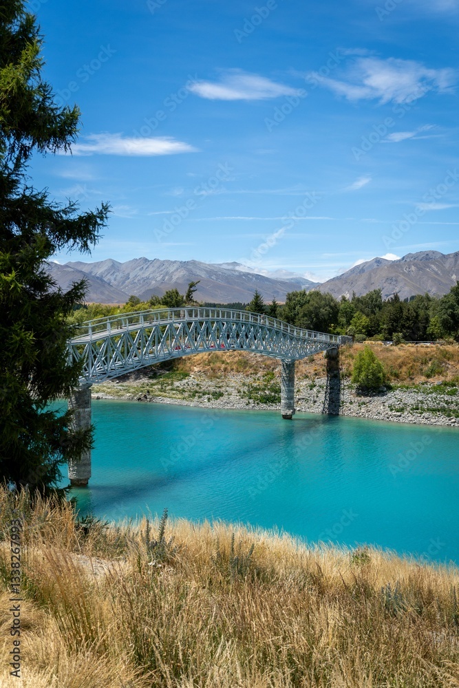 Obraz premium A scenic view of a turquoise river with a metal bridge and mountains at Tekapo, New Zealand.