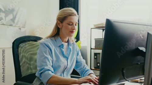 Charming Caucasian female sitting at desk and typing on keyboard. Smiling slightly while working on computer. Filling out information in PC. Bright modern office with shelves and decorative elements.