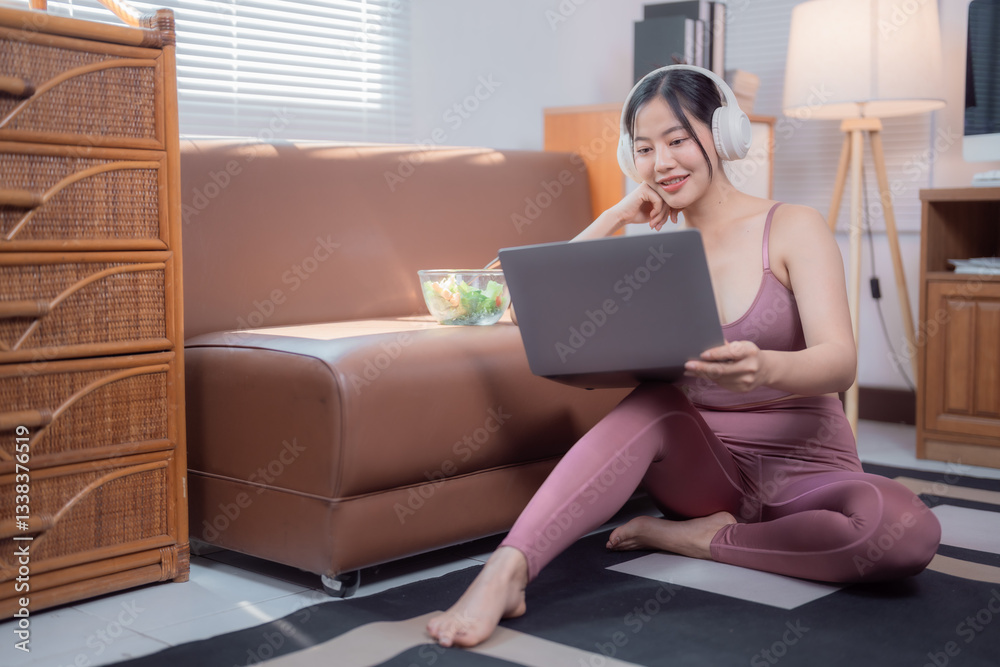 Fototapeta premium Young sportswoman wearing headphones using laptop and eating salad at home, sitting on the floor with a bowl of salad on the sofa next to her, she's enjoying healthy lifestyle