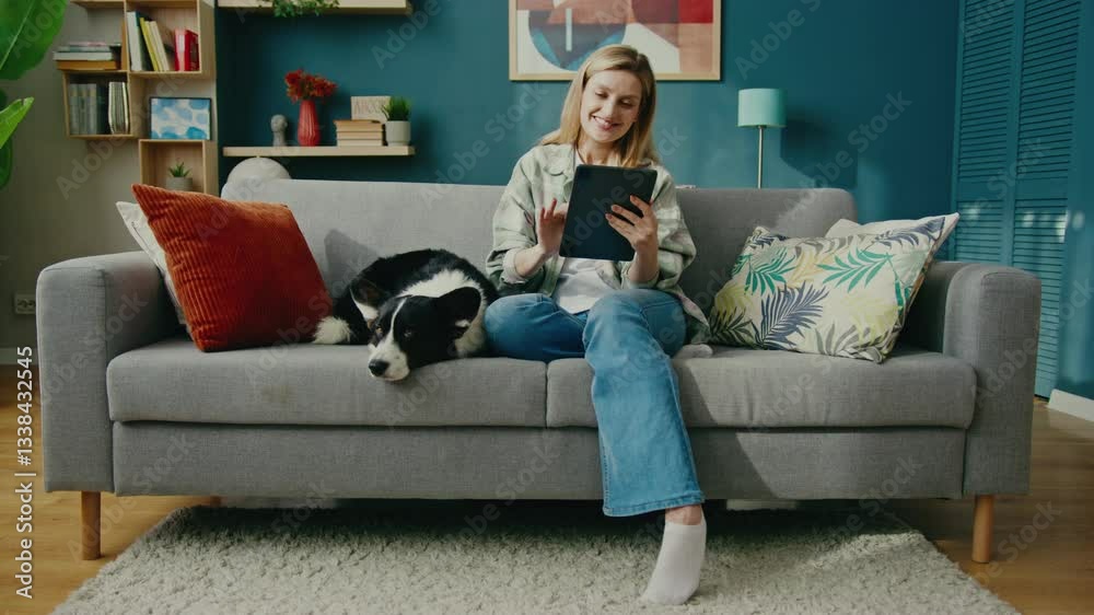 Smiling Caucasian female sitting on big sofa and holding tablet while gently stroking relaxed corgi. Enjoying peaceful moment while reading or watching content in bright stylish living room. Smiling Caucasian female sitting on big sofa and holding tablet while gently stroking relaxed corgi. Enjoying peaceful moment while reading or watching content in bright stylish living room.