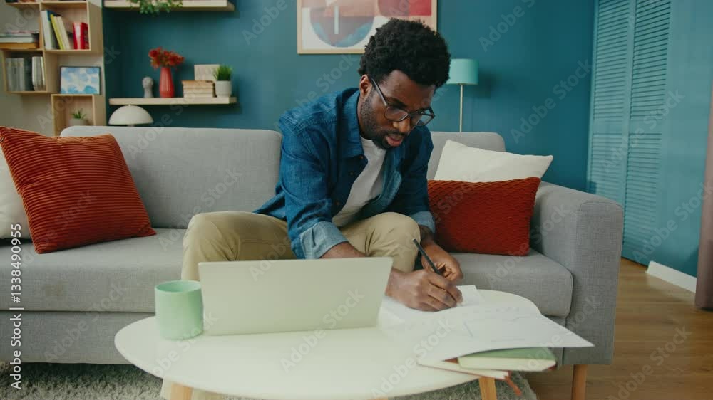 African American man in glasses analyzing financial chart at home. Focused entrepreneur or freelancer reviewing business data. Taking notes on investment strategy. Remote work and planning concept. African American man in glasses analyzing financial chart at home. Focused entrepreneur or freelancer reviewing business data. Taking notes on investment strategy. Remote work and planning concept.
