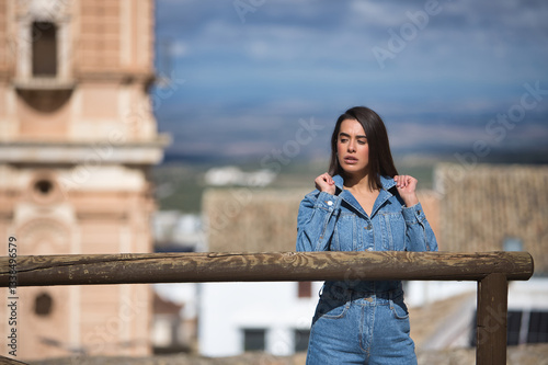 Young, beautiful, brunette, Hispanic woman, wearing a denim suit, raising her shirt collars in a cocky attitude, with an urban landscape in the background. Concept fashion, beauty, trendy, femininity.