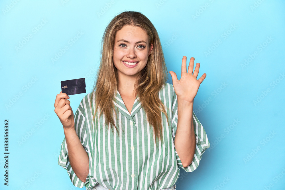 Young blonde with a credit card in blue studio smiling cheerful showing number five with fingers. Young blonde with a credit card in blue studio smiling cheerful showing number five with fingers.
