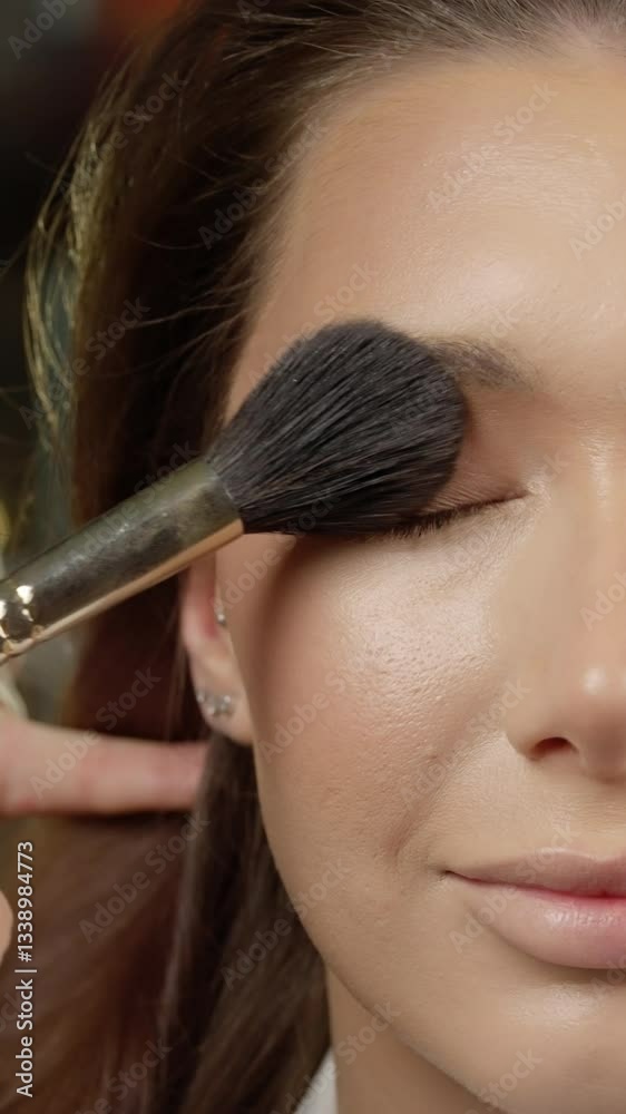 Professional makeup artist blending foundation with precision brush, creating flawless complexion in minimalist studio environment with soft lighting