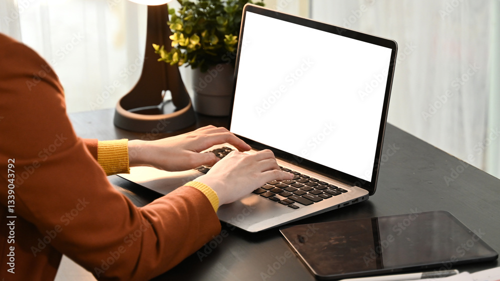 A woman is typing on a laptop with a white screen. The laptop is on a table next to a tablet.