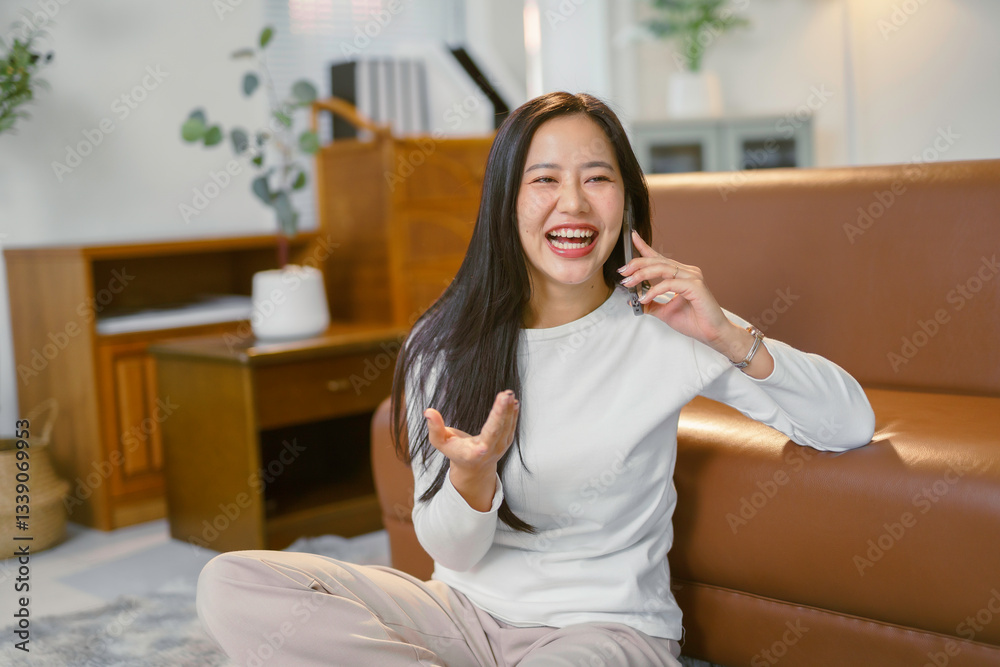 Cheerful young woman sitting on the floor by the sofa, talking on mobile phone and laughing, enjoying conversation Cheerful young woman sitting on the floor by the sofa, talking on mobile phone and laughing, enjoying conversation