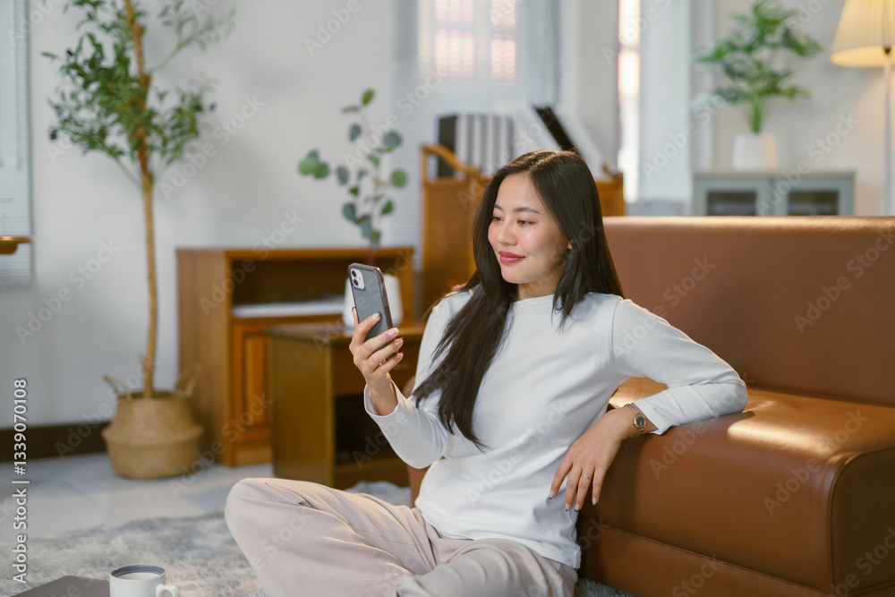 Young woman relaxing cross legged near sofa, scrolling smartphone and messaging friends in cozy living space Young woman relaxing cross legged near sofa, scrolling smartphone and messaging friends in cozy living space