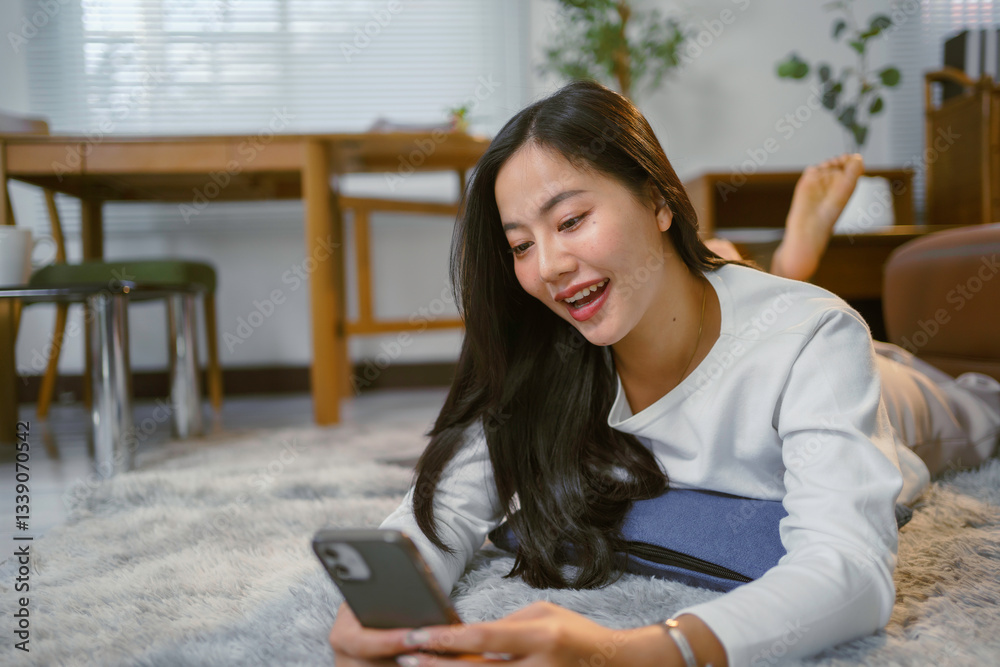 Smiling Asian woman lounging on soft carpet, browsing smartphone while relaxing in comfortable home setting Smiling Asian woman lounging on soft carpet, browsing smartphone while relaxing in comfortable home setting