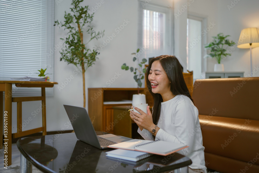 Cheerful freelancer working remotely, videoconferencing on laptop while drinking coffee in stylishly decorated home office space Cheerful freelancer working remotely, videoconferencing on laptop while drinking coffee in stylishly decorated home office space