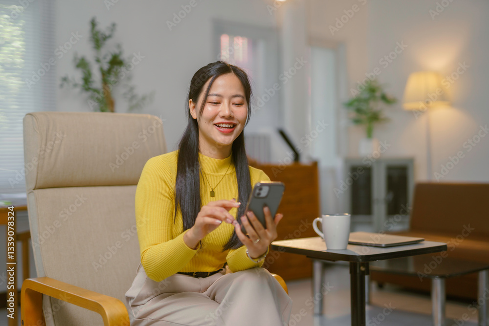 Smiling young Asian businesswoman using smartphone while sitting on armchair at home office Smiling young Asian businesswoman using smartphone while sitting on armchair at home office