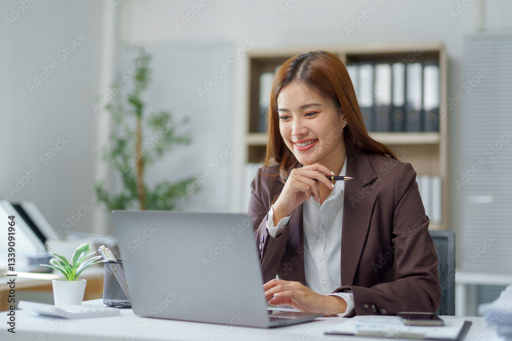 Professional Asian female concentrating while typing on laptop, pen in hand, radiating confidence in contemporary workspace Professional Asian female concentrating while typing on laptop, pen in hand, radiating confidence in contemporary workspace