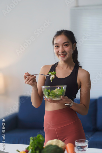 Happy asian sportswoman eating a bowl of fresh salad in her modern apartment after training