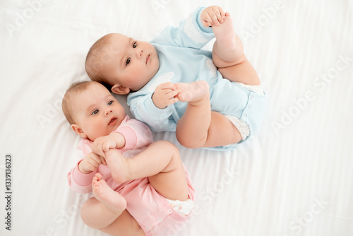 Two babies, a boy and a girl, on a white bed in a bedroom, lying on the back, a cute little child of six months playing with his legs, a place for text, top view