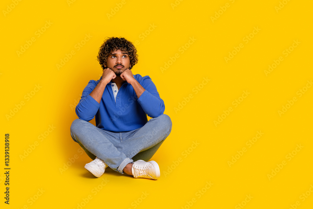 Young man sitting against a yellow background wearing a blue polo shirt and jeans, depicting casual fashion and thoughtful leisure Young man sitting against a yellow background wearing a blue polo shirt and jeans, depicting casual fashion and thoughtful leisure