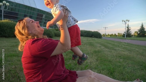 A young woman is sitting on the grass in the park in the summer, her little son is running towards her. The mother takes the child in her arms and kisses him. Entertainment with a child and a mother