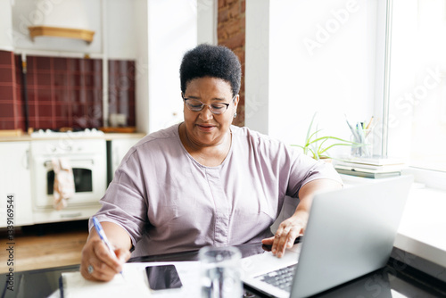 Portrait of elderly black plus size female studying at home sitting at kitchen table in front of laptop next to window, watching webinar, noting down tips and information after speaker