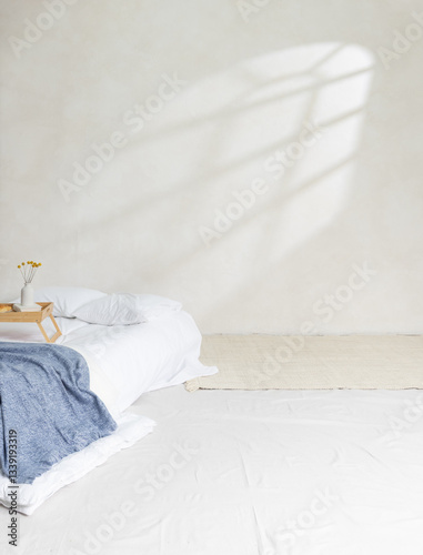 A minimalist bedroom scene featuring a low-profile bed with white linens and a soft blue blanket, placed against a neutral-toned textured wall. Sunlight filters through an unseen window