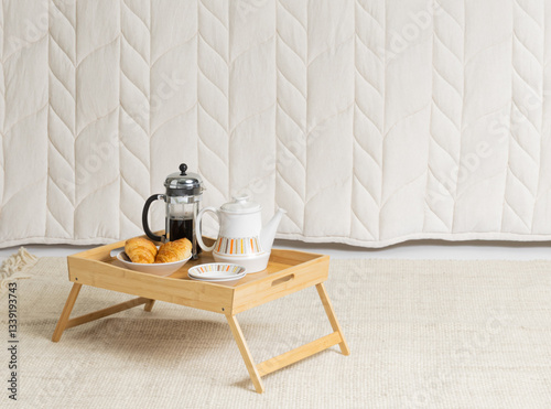 A cozy indoor scene featuring a light bamboo folding breakfast tray with a French press, teapot, and croissants, set against a textured off-white quilted backdrop and a neutral woven rug, evoking a re