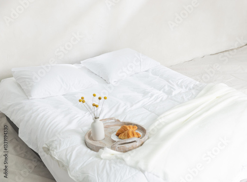 A minimalist, bright, and airy bedroom scene featuring a white duvet with two pillows, a wooden tray with two croissants and a small vase of yellow billy balls, set against a simple white wall