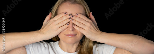 Glad smiling blonde female model hiding her face behind hand, has broad smile, happy to recieve compliments from handsome man, isolated against black background. Positive young woman