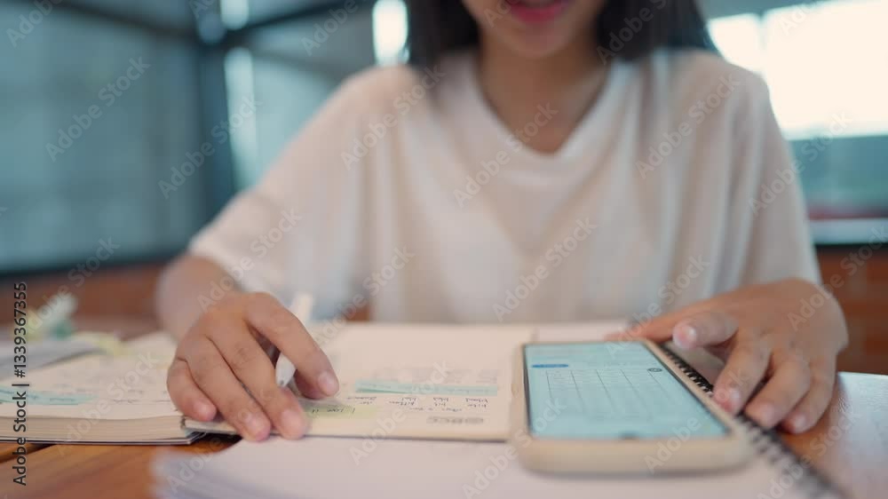 A woman is writing in a notebook while looking at her phone. She is focused on her work and seems to be multitasking A woman is writing in a notebook while looking at her phone. She is focused on her work and seems to be multitasking