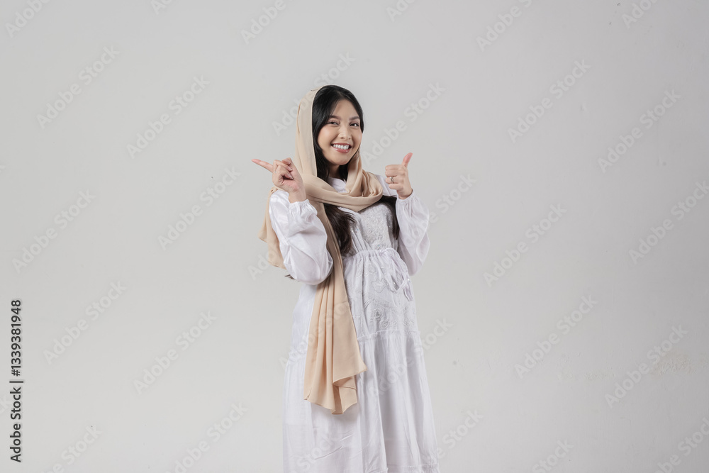 Asian Muslim pregnant woman pointing to the copy space behind her while giving thumb up gesture. Asian Muslim pregnant woman pointing to the copy space behind her while giving thumb up gesture.