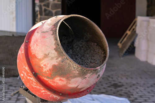 Red concrete mixer with wet cement inside at construction site. Close-up shot of rotating drum covered in dried cement. Industrial construction and building process concept.